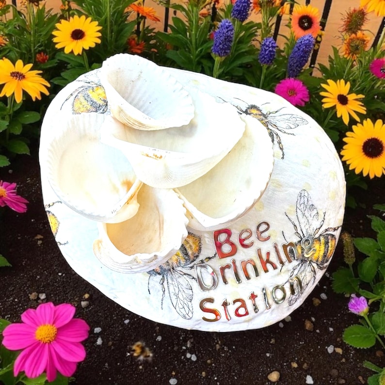 May include: A white, shell-shaped bee drinking station with multiple levels. The station is decorated with painted bees and the words "Bee Drinking Station" in red and silver lettering. Surrounded by colorful flowers.