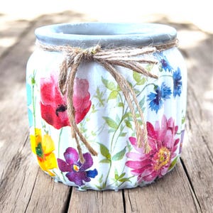 May include: A white, cylindrical jar decorated with colourful floral patterns, including red poppies, purple pansies, and blue cornflowers. The jar has a grey rim and a twine bow, suitable for home decor or as a gift.