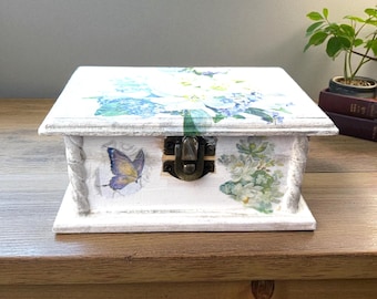 Puede incluir: Una caja de madera blanca con tapa con bisagras, decorada con calcomanías florales y de mariposas. La caja tiene un cierre de metal y un acabado envejecido. Es un artículo de almacenamiento decorativo.