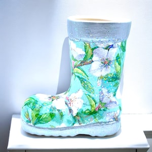 May include: A blue ceramic boot planter with a white glitter finish. The boot is decorated with a floral pattern of white flowers and green leaves.