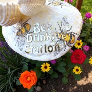 May include: A white, oval-shaped bee drinking station with the words "Bee Drinking Station" in silver letters. The station is decorated with illustrations of bees and surrounded by colorful flowers, including orange and red roses, and yellow and pink daisies.