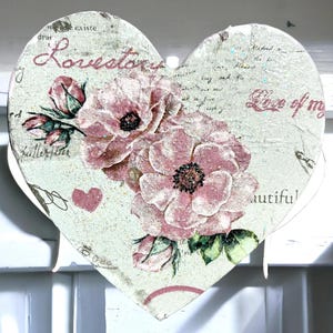 A heart-shaped decoration features a textured, glittery surface adorned with an intricate floral design. Soft pink blossoms intertwine with green leaves, creating a delicate, vintage aesthetic. Scattered text in varied fonts, including "Lovetown" and "Love of my," overlays the pattern, blending into the composition. The close-up reveals fine details, such as subtle shading and the embossed texture, enhancing the tactile and visual depth of the piece