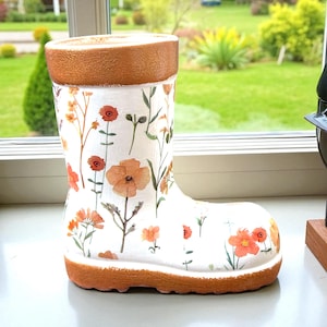 Puede incluir: Una maceta con forma de bota de cerámica blanca con un diseño floral con flores naranjas y rojas y hojas verdes. La parte superior e inferior de la bota son de color marrón cálido. La bota está en un alféizar.