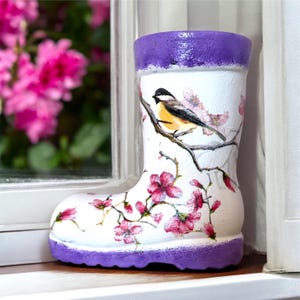 May include: A white boot-shaped planter with a purple rim and base. The surface features a painted design of a small bird perched on a brown branch, surrounded by pink cherry blossom flowers. The finish appears textured and hand-painted.