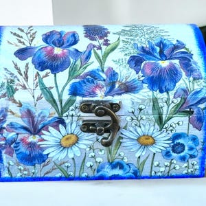 Op de afbeelding: Een houten doos met een scharnierend deksel, versierd met een bloemenpatroon van blauwe irissen en witte madeliefjes. De doos heeft een messing sluiting en een blauwe rand rond de randen.