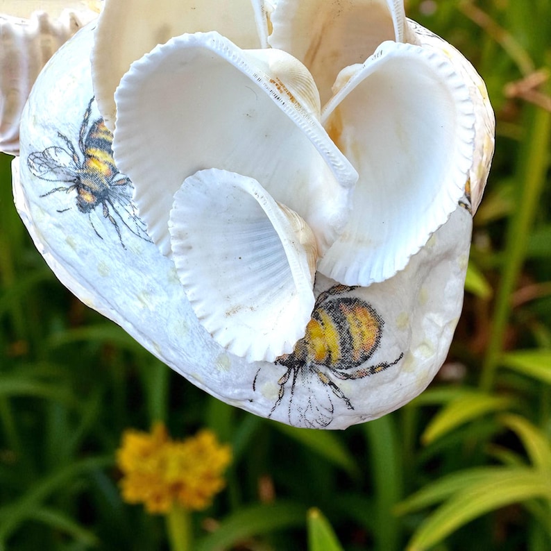 May include: A decorative heart-shaped ornament crafted from white seashells. The ornament features printed bee illustrations in yellow, black, and gray. The seashells are arranged to create a layered effect, with a blurred green background.