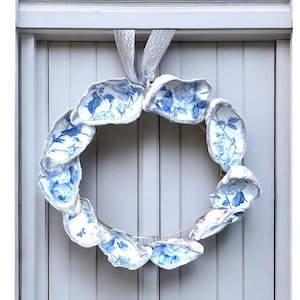 Peut inclure: Une couronne décorative avec un cadre métallique circulaire orné de coquillages à motifs floraux blancs et bleus. Les coquillages sont disposés en cercle et sont bordés d'une garniture argentée scintillante. Un ruban argenté est utilisé pour la suspension.
