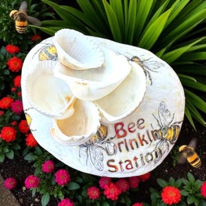 Puede incluir: Una estación de abrevadero para abejas en forma de concha blanca, con múltiples cuencos de agua. La estación está decorada con ilustraciones de abejas y las palabras "Bee Drinking Station" en letras rojas y plateadas. Rodeada de flores rojas y rosas y follaje verde.