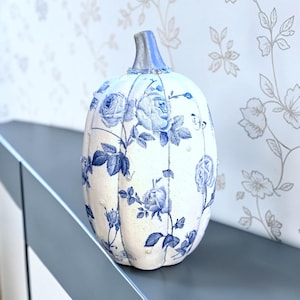 May include: A decorative pumpkin featuring a white base with a blue floral pattern of roses and leaves. The pumpkin has a silver-painted stem and is accented with small, scattered white pearls along its ribbed surface and around the stem base.