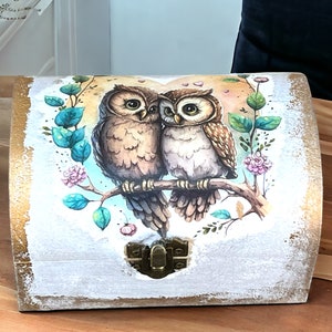 Peut inclure: Une boîte en bois avec une bordure dorée et un motif peint de deux hiboux perchés sur une branche avec des fleurs roses. La boîte a une ouverture en forme de cœur et un loquet en métal.