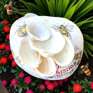 May include: A white, round, decorative shell planter with a bee theme. The planter is adorned with illustrations of bees and has several large, white shells attached. The planter is surrounded by red and pink flowers and green foliage.