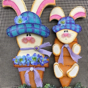 May include: Two wooden Easter bunny decorations. One bunny is holding a carrot and wearing a blue and white checked hat. The other bunny is in a flower pot and wearing a blue and white checked hat with a purple ribbon.
