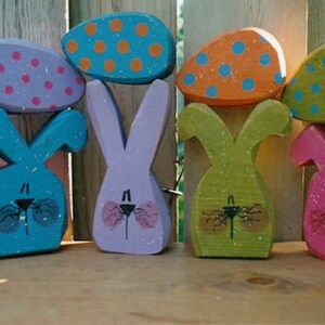 May include: Five wooden bunny figurines with painted details. Each bunny is topped with a different colored egg with polka dots. The eggs are pink, orange, green, orange, and pink.