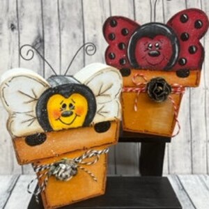 May include: Two wooden planter boxes shaped like a bee and a ladybug. The bee planter is yellow with black stripes and white wings. The ladybug planter is red with black spots and black wings. Both planters have brown pots with a pine cone and twine decoration.