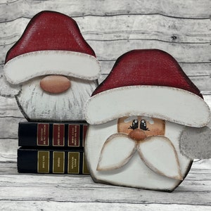 May include: Two wooden Santa Claus figures with red hats, white beards, and painted faces. The figures are standing on a stack of books with gold lettering.