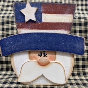 May include: Wooden cutout of a face with a white beard and moustache, wearing a blue and white hat with a red, white and blue American flag on top.