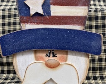May include: Wooden cutout of a face with a white beard and moustache, wearing a blue and white hat with a red, white and blue American flag on top.