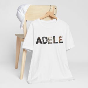May include: White t-shirt with the name "ADELE" printed across the front. The letters are filled with images of the singer. The shirt is hanging on a wooden hanger attached to a light-colored wooden chair. A white baseball cap rests on the chair.