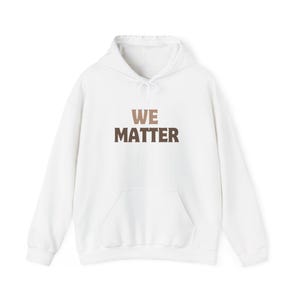 Op de afbeelding: Witte hoodie met een voorzak en een trekkoord. De woorden "WE MATTER" zijn in bruine en bruine blokletters op de borst gedrukt. De hoodie is gemaakt van een zacht materiaal.