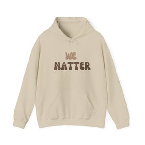 Puede incluir: Sudadera con capucha color crema con bolsillo delantero y capucha. El texto "WE MATTER" está impreso en la parte delantera en letras marrones y beige. La sudadera está hecha de un material suave.
