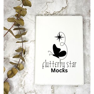 May include: A white notebook with the text "flutterby star Mocks" and a black butterfly and star design. The notebook is on a marble surface with dried eucalyptus leaves.