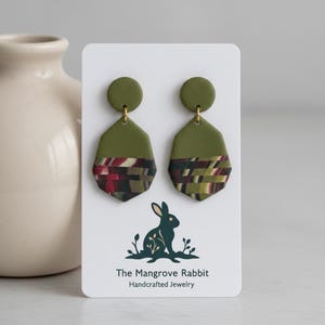 May include: A pair of olive green dangle earrings featuring a circular top and a hexagonal bottom section with a multi-colored striped pattern in shades of red, black, and green. The earrings are displayed on a white card with a rabbit logo and the text The Mangrove Rabbit Handcrafted Jewelry.