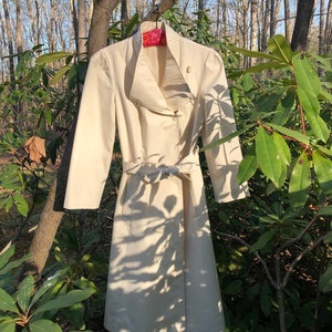 May include: A white trench coat with a wide collar and a belt. The coat is hanging on a hanger in front of a green leafy background.