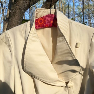 May include: A white jacket with a large, folded collar and a single button closure. The jacket is hanging on a floral patterned hanger.