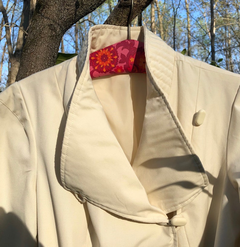 May include: A white jacket with a large, folded collar and a single button closure. The jacket is hanging on a floral patterned hanger.