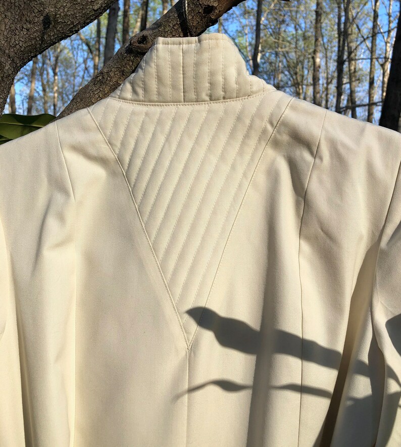 May include: A cream-colored jacket with a quilted back panel. The quilting is in a diamond pattern and the jacket has a high collar.