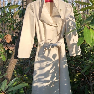 May include: A white trench coat with a large collar, a belt, and a button closure. The coat is hanging on a hanger in front of a green leafy background.