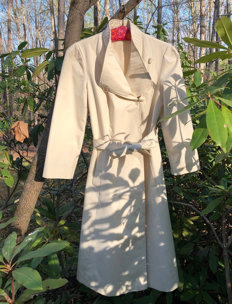 May include: A white trench coat with a large collar, a belt, and a button closure. The coat is hanging on a hanger in front of a green leafy background.