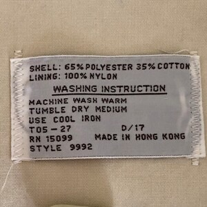 May include: A white tag with black text detailing the fabric composition and washing instructions for a garment. The tag reads "SHELL: 65% POLYESTER 35% COTTON LINING: 100% NYLON WASHING INSTRUCTION MACHINE WASH WARM TUMBLE DRY MEDIUM USE COOL IRON T05-27 RN 15099 D/17 MADE IN HONG KONG STYLE 9992".