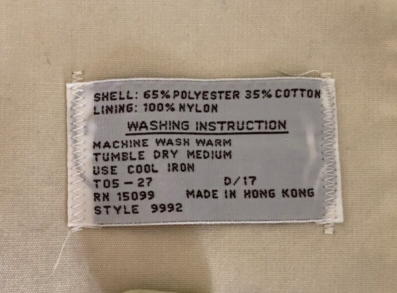 May include: A white tag with black text detailing the fabric composition and washing instructions for a garment. The tag reads "SHELL: 65% POLYESTER 35% COTTON LINING: 100% NYLON WASHING INSTRUCTION MACHINE WASH WARM TUMBLE DRY MEDIUM USE COOL IRON T05-27 RN 15099 D/17 MADE IN HONG KONG STYLE 9992".