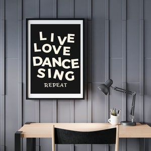 May include: A framed black and white print with the words "LIVE LOVE DANCE SING REPEAT" in a playful font. The print hangs above a wooden desk with a black lamp, a white mug with pencils, and a black chair.