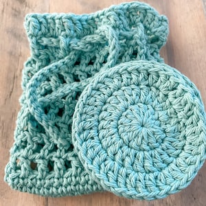 May include: A crocheted set in a light turquoise color. The set includes a round face scrubby and a drawstring bag. The bag has an open weave design and a braided handle. The scrubby is circular with a textured surface.