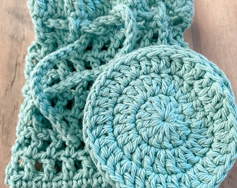 May include: A light turquoise crocheted set featuring a round face scrubby and a drawstring bag. The bag has an open weave design and a braided handle. The scrubby is circular with a textured surface, perfect for skincare.