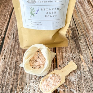 May include: A kraft paper bag labeled "Caroline's Homemade Home" contains "Relaxing Bath Salts" with lavender and cedarwood. A small muslin bag and wooden scoop hold pink bath salts, with some spilled on the wooden surface. The bag contains 300 g of bath salts.