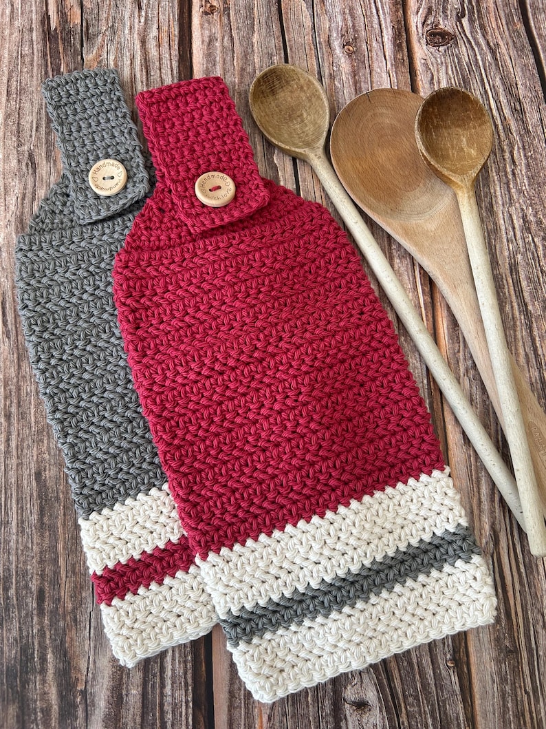 Op de afbeelding: Twee handgemaakte gehaakte keukendoeken in grijs en rood, met een gestreepte onderkant in wit, grijs en rood. Elke handdoek heeft een houten knoopsluiting. Houten lepels op de achtergrond.