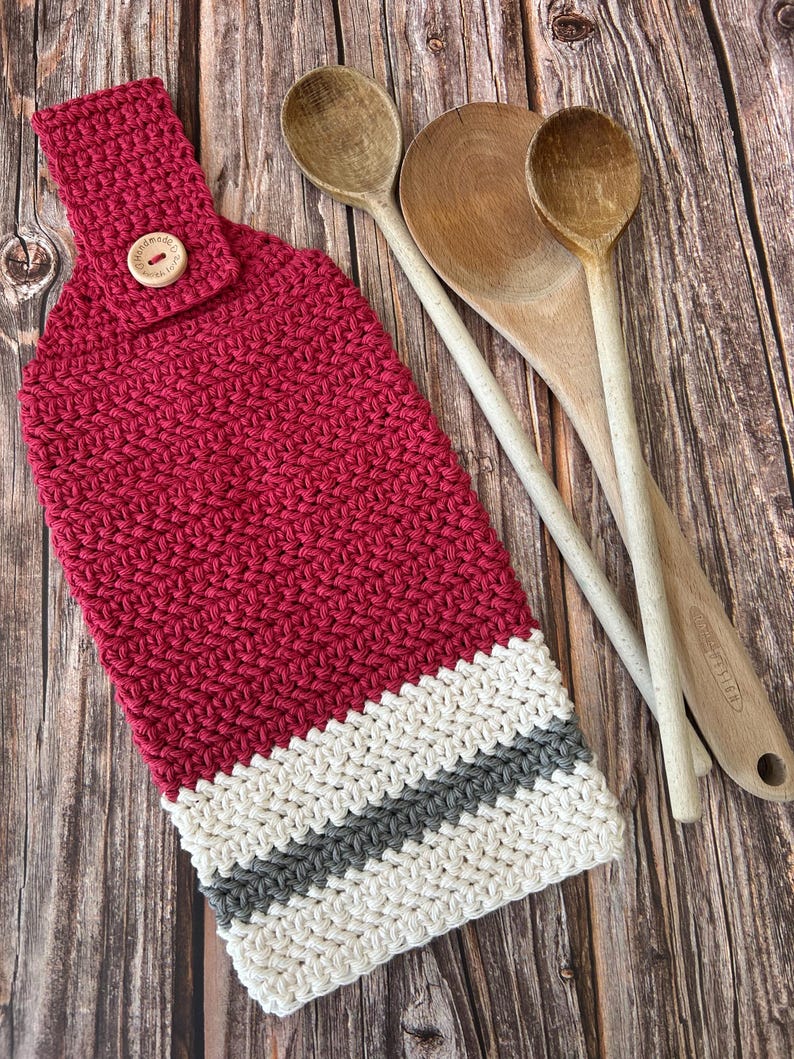 Op de afbeelding: Een handgemaakte, rode gehaakte keukendoek met een wit en grijs gestreepte onderkant. De handdoek heeft een lus met een houten knoop om op te hangen. Drie houten lepels liggen naast de handdoek op een houten oppervlak.