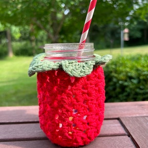 Puede incluir: Un tarro de cristal transparente con una funda de fresa de ganchillo roja y un borde de hoja de ganchillo verde. Una pajita a rayas rojas y blancas está insertada en el tarro. El tarro está sobre una superficie de madera marrón.