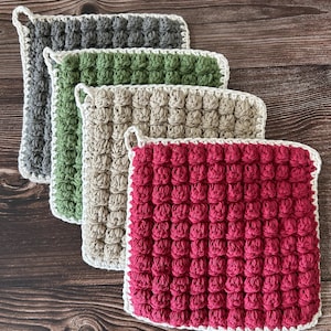 Puede incluir: Cuatro agarraderas de ganchillo hechas a mano en tonos de gris, verde, beige y rojo. Cada agarradera cuadrada presenta un patrón de puntada en relieve texturizado y un borde blanco con un lazo para colgar. Las agarraderas están apiladas.