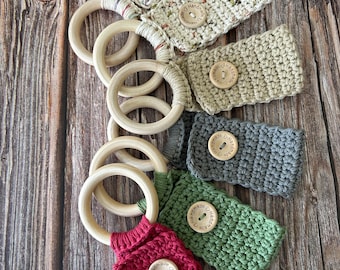 Puede incluir: Una colección de anillos de juguete para bebés de ganchillo. Los anillos de madera clara están unidos a fundas de ganchillo en varios colores, incluyendo rosa, verde y gris. Cada funda tiene un botón de madera con la inscripción "Handmade".