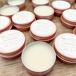 May include: A close-up of several small, rose gold tins filled with lip balm. The tins have white labels with the text "Caroline's Homemade Home" and "Peppermint Lip Balm 20 ml".