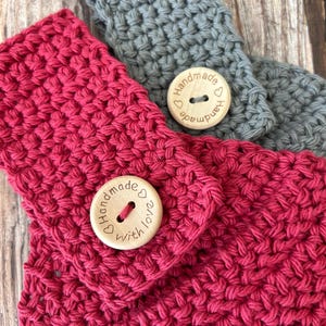 Op de afbeelding: Close-up van twee handgemaakte gehaakte hoofdbanden. E&eacute;n is fel fuchsia, de andere is grijs. Elke hoofdband heeft een houten knoop met de woorden "Handmade with love" gegraveerd. De haaksteek cre&euml;ert een textuur.