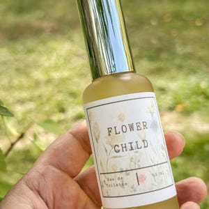 Peut inclure: Un flacon de parfum avec un bouchon argenté et une étiquette blanche. L'étiquette indique "FLOWER CHILD" avec des illustrations florales et "Eau de Toilette" et "50 ml" imprimés en dessous. Le flacon contient un liquide jaune.