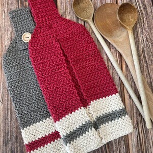 Op de afbeelding: Twee handgemaakte gehaakte keukendoeken. E&eacute;n is grijs met een knoopsluiting en de andere is rood. Beide handdoeken hebben een gestreept ontwerp aan de onderkant met witte en grijze strepen. Houten lepels staan op de achtergrond.
