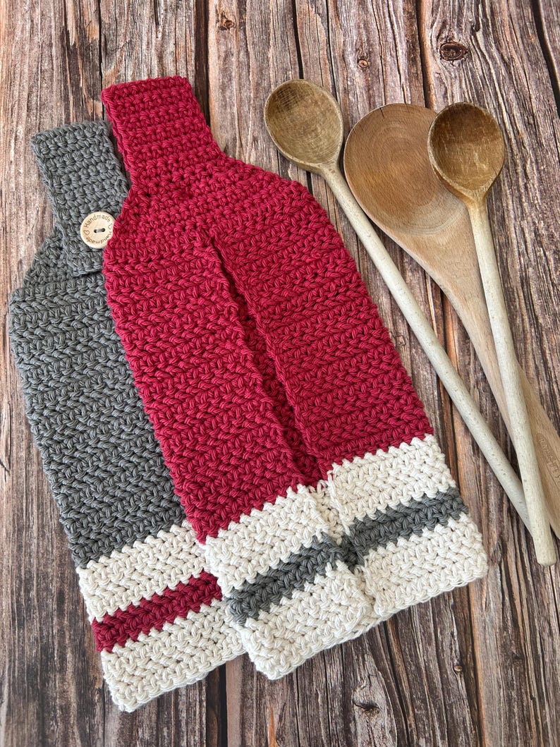 Op de afbeelding: Twee handgemaakte gehaakte keukendoeken. E&eacute;n is grijs met een knoopsluiting en de andere is rood. Beide handdoeken hebben een gestreept ontwerp aan de onderkant met witte en grijze strepen. Houten lepels staan op de achtergrond.