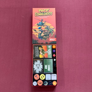 May include: A board game box set titled "Dice Commandos" with a red and orange color scheme. The box features illustrations of soldiers and a helicopter. Inside, there are cards, dice, and tokens. The game is designed for solo play.