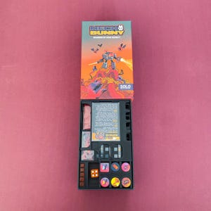 May include: An open board game box with the title "MECH BUNNY" and a colorful illustration of a robot on a mountain. The box contains game pieces, dice, and cards. The game is designed by Gabe Barrett.